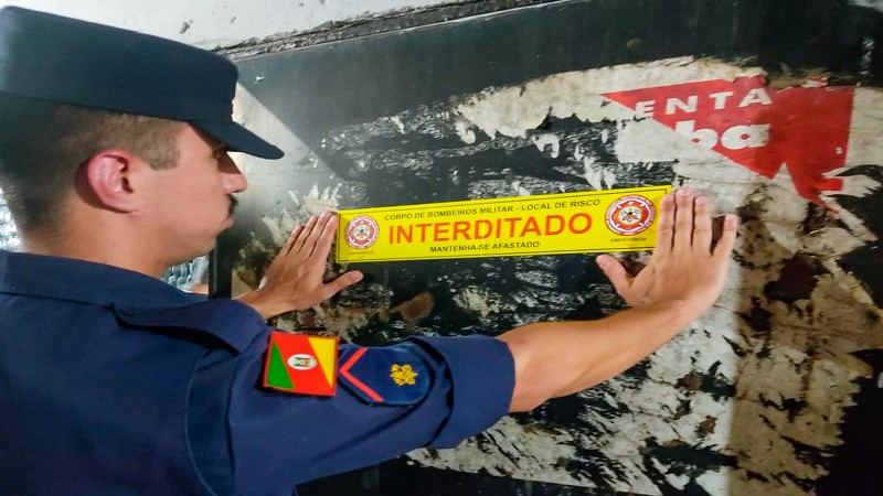 Na imagem, bombeiro militar fixa adesivo na parede escrito interditado.