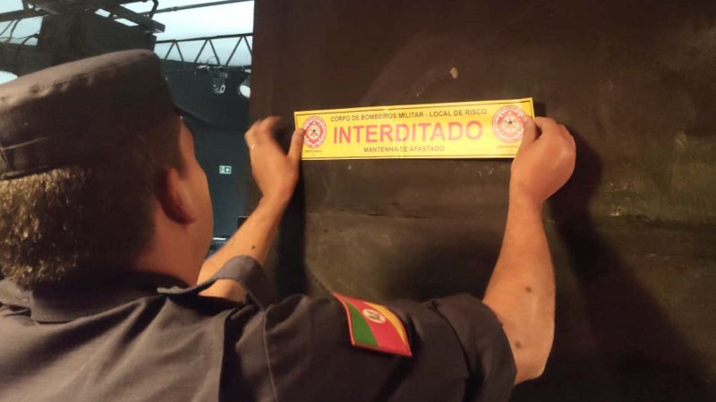 Na foto, bombeiro militar fixa adesivo na parede escrito: CORPO DE BOMBEIROS MILITAR - LOCAL DE RISCO - INTERDITADO - MANTENHA-SE AFASTADO.